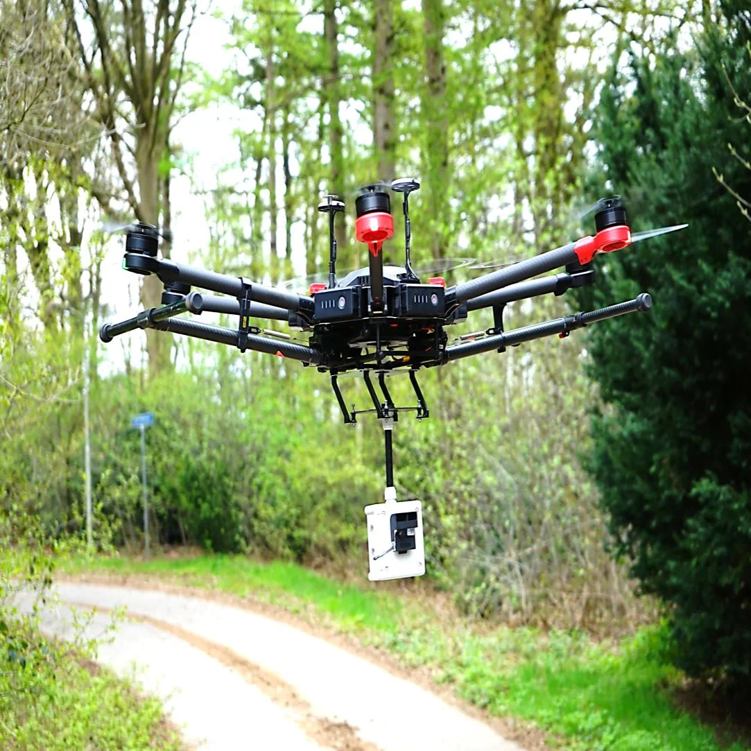 Quadcopter drone in flight over a woodland path, carrying a white sensor box beneath it